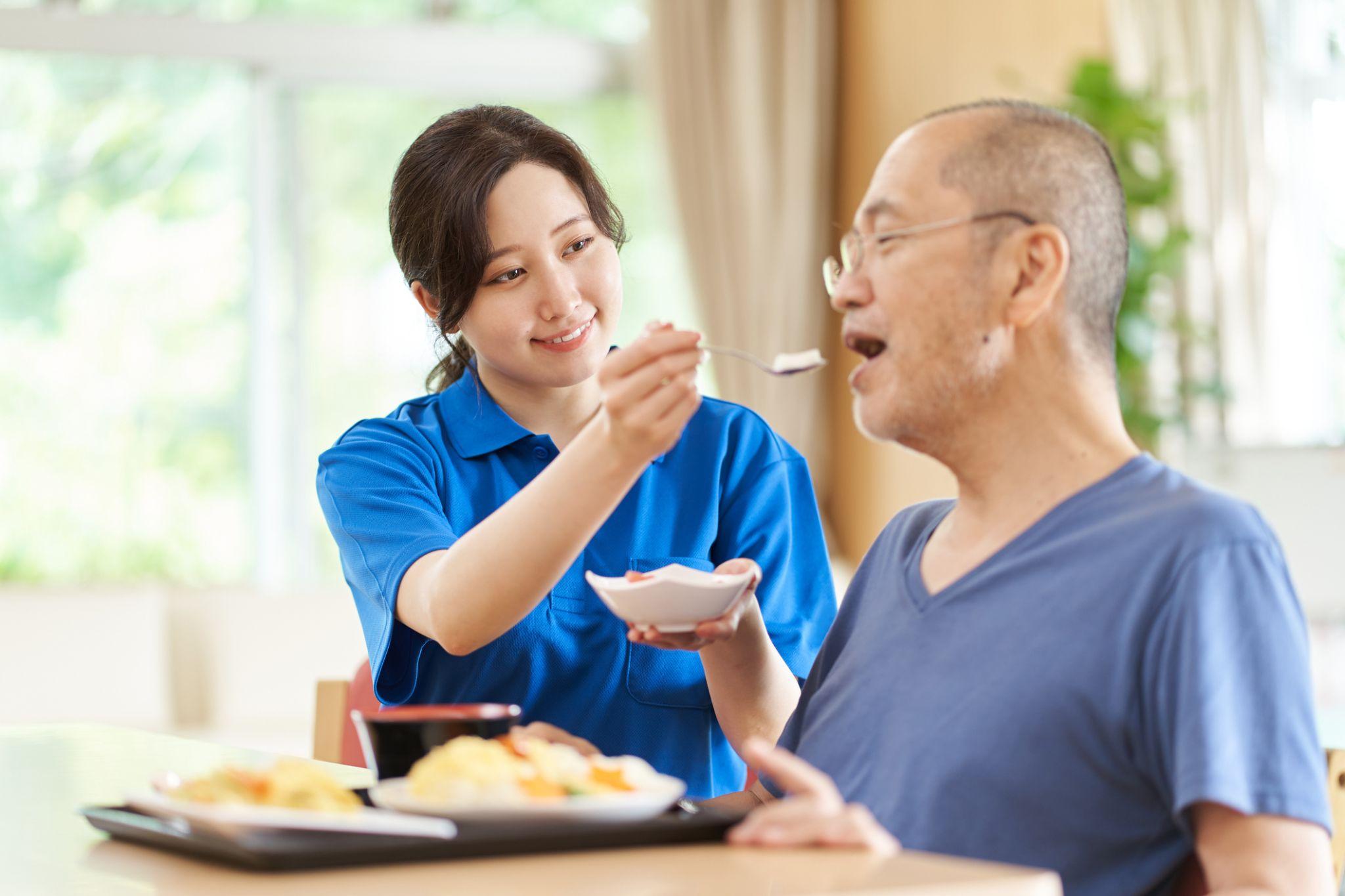 female aid in blue shirt feeds senior man in blue shirt wearing glasses in his home