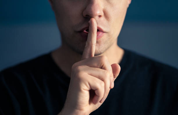 A caucasian man holds his index finger to his lips to nonverbally communicate that silence is needed.