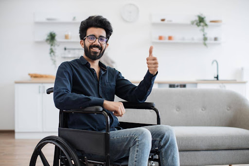 A disabled man in a wheelchair sitting in a living area smiling and making a thumbs-up sign.