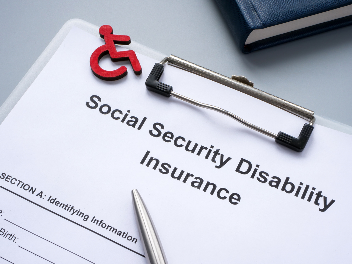 An SSDI form on a clipboard with a pen and a small red model of a person in a wheelchair sitting on top of it.